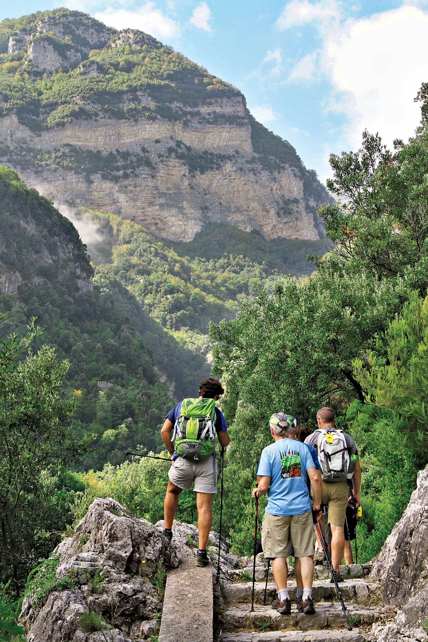 A group of hikers traversing a rocky trail through a lush, mountainous landscape with towering cliffs in the background.