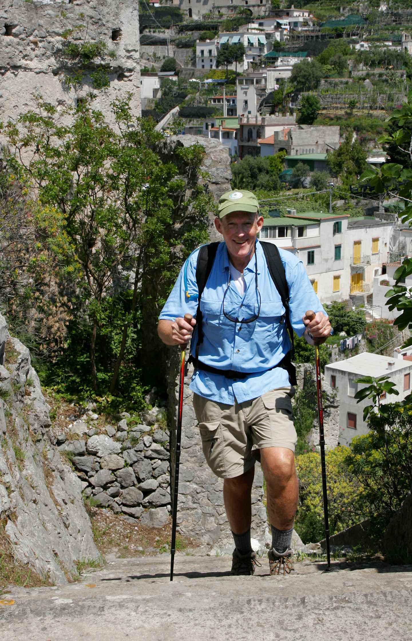An elderly man in hiking gear is walking up a set of stone steps, with a scenic village nestled in the background among lush greenery.