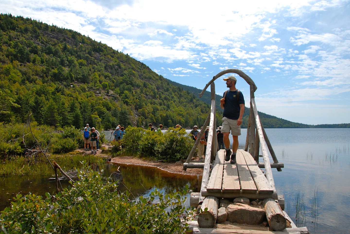 A wooden bridge extends across a serene lake, surrounded by lush green forests and mountains in the background, with several people walking along the bridge.