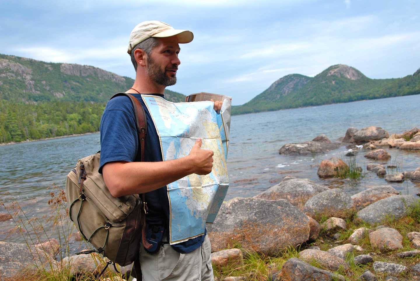 A man in casual attire stands on a rocky shore, holding a map and gazing out at the scenic landscape of mountains and a lake in the background.
