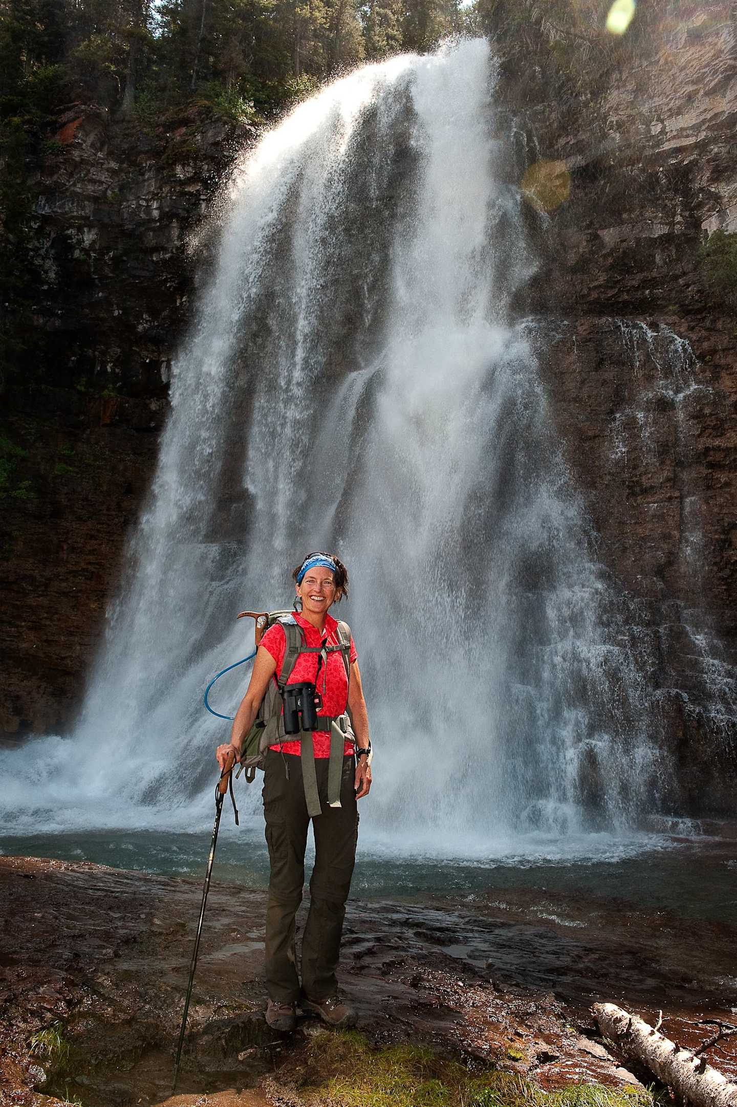 A person in a red life jacket and hiking gear stands in the foreground, facing a large, cascading waterfall in a lush, forested environment.