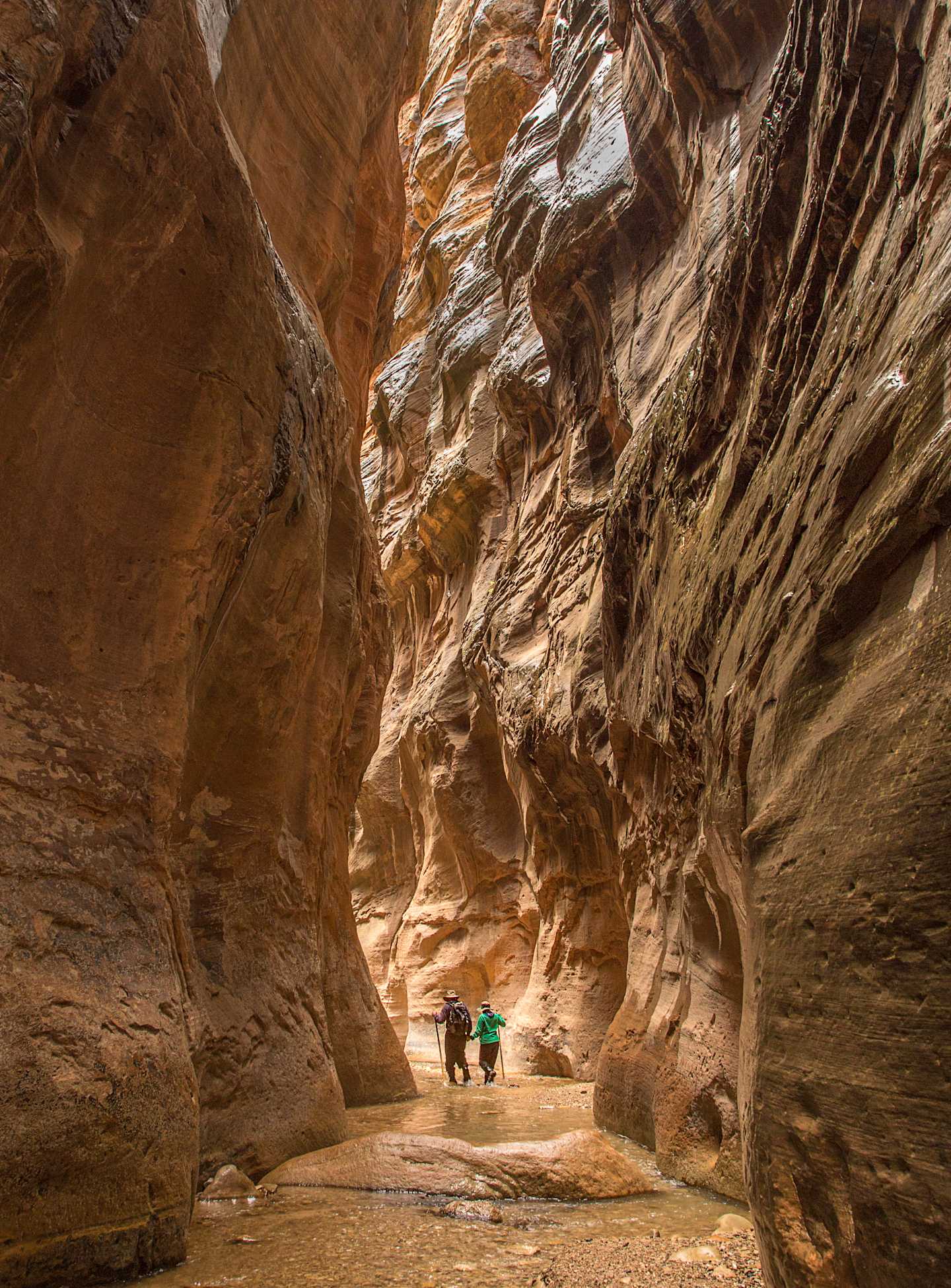 A narrow, winding canyon with towering, rugged sandstone walls surrounds two hikers in the foreground, creating an awe-inspiring and dramatic natural landscape.