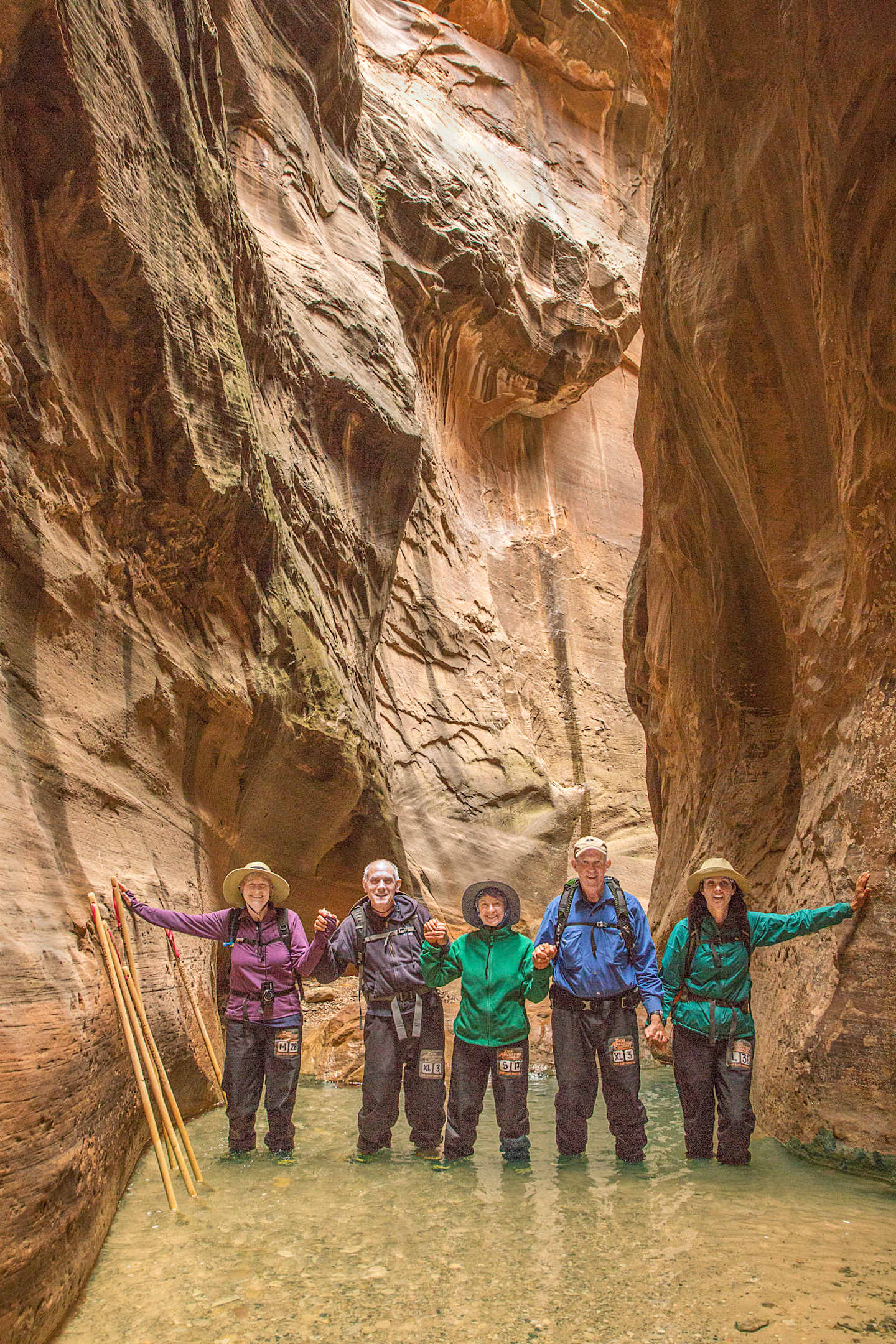 A group of hikers standing in a shallow stream surrounded by towering, multicolored rock formations in a narrow canyon.