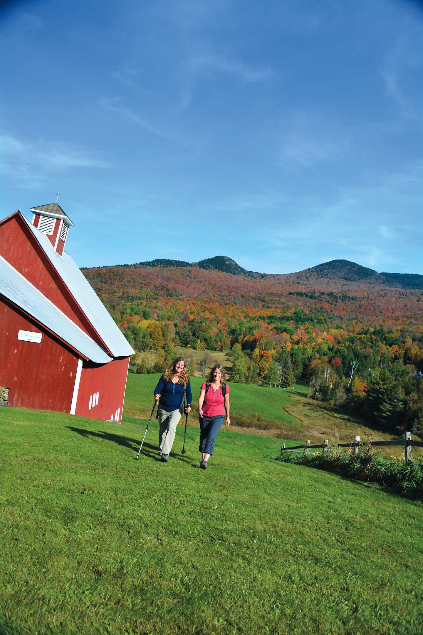 A red barn stands in the foreground, with a couple walking on the grassy field in front of it. In the background, a scenic autumn landscape with colorful trees and rolling hills can be seen under a clear blue sky.