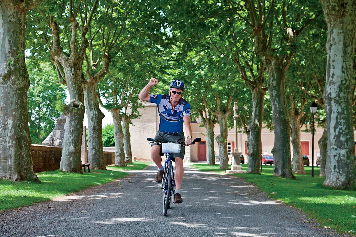 A person on a bicycle rides down a tree-lined path surrounded by lush greenery in a park-like setting.