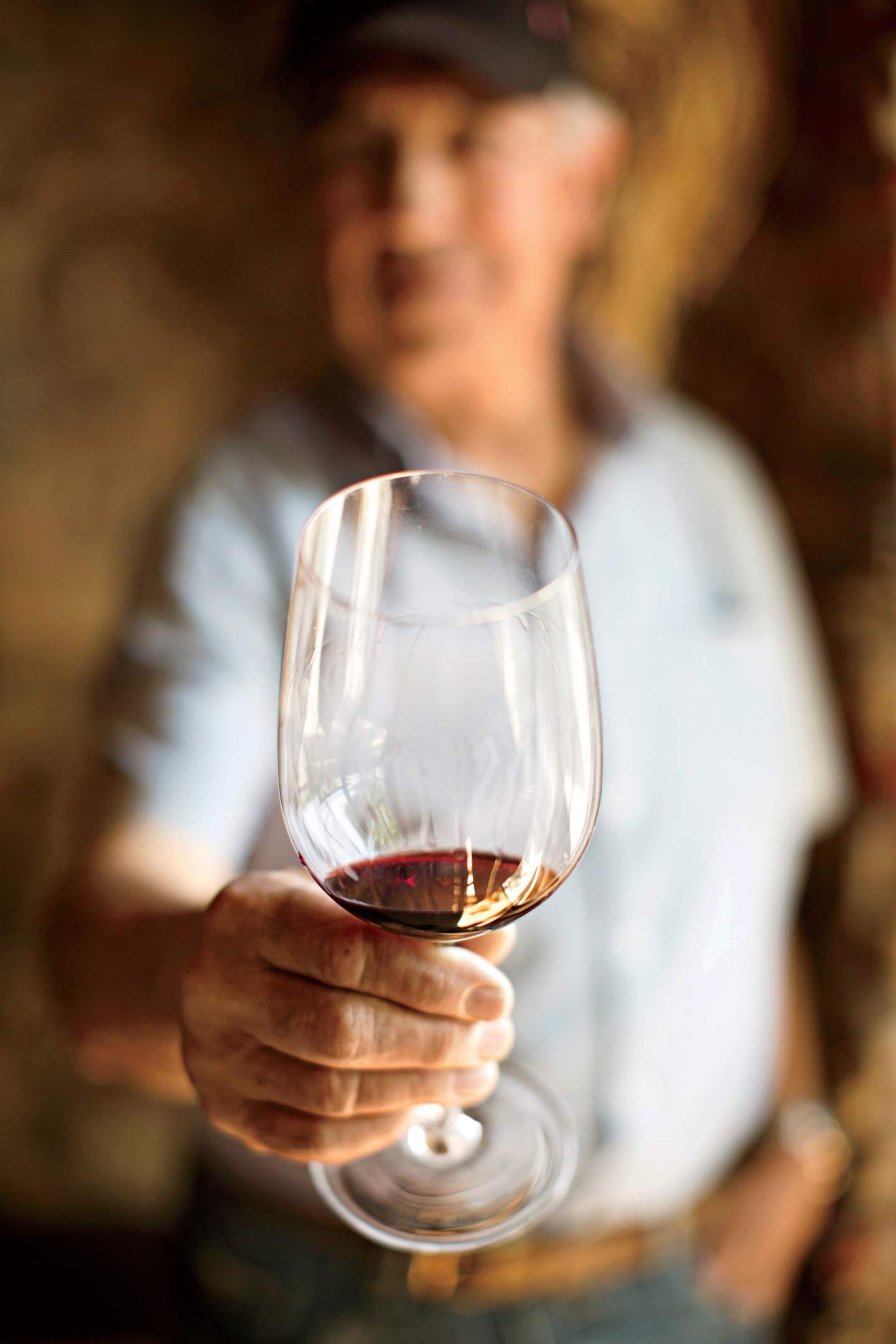 A person in a white shirt and hat is holding a glass of red wine against a blurred background.