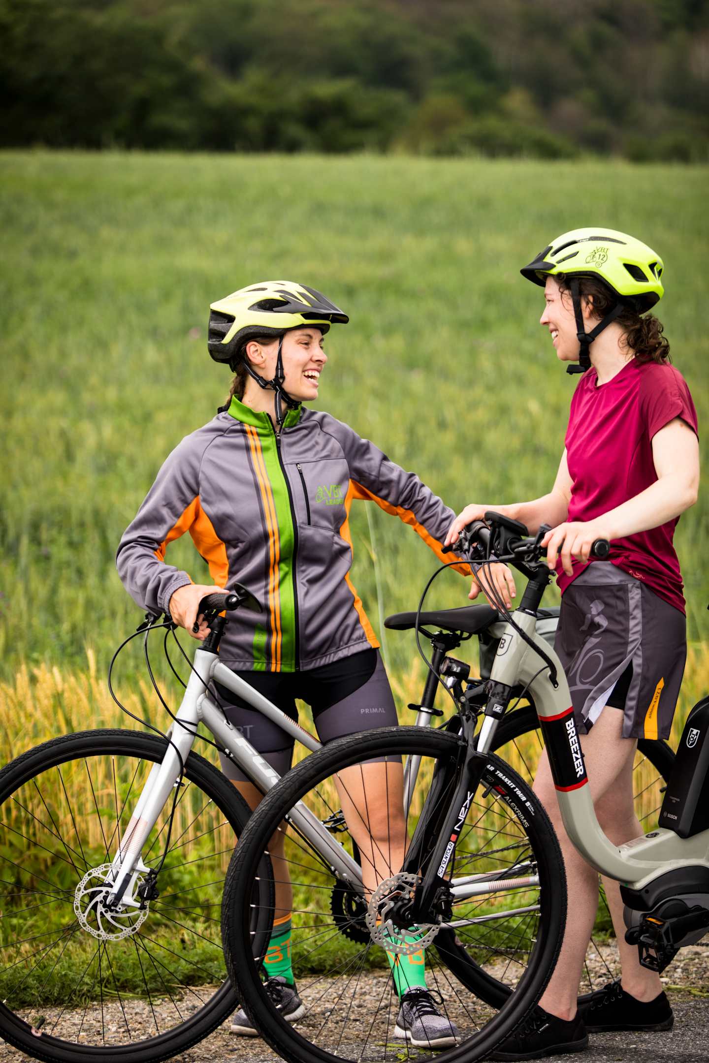 Two cyclists, a woman in a gray and neon jacket and a woman in a red shirt, are riding their bicycles on a dirt path surrounded by a grassy field and trees in the background.