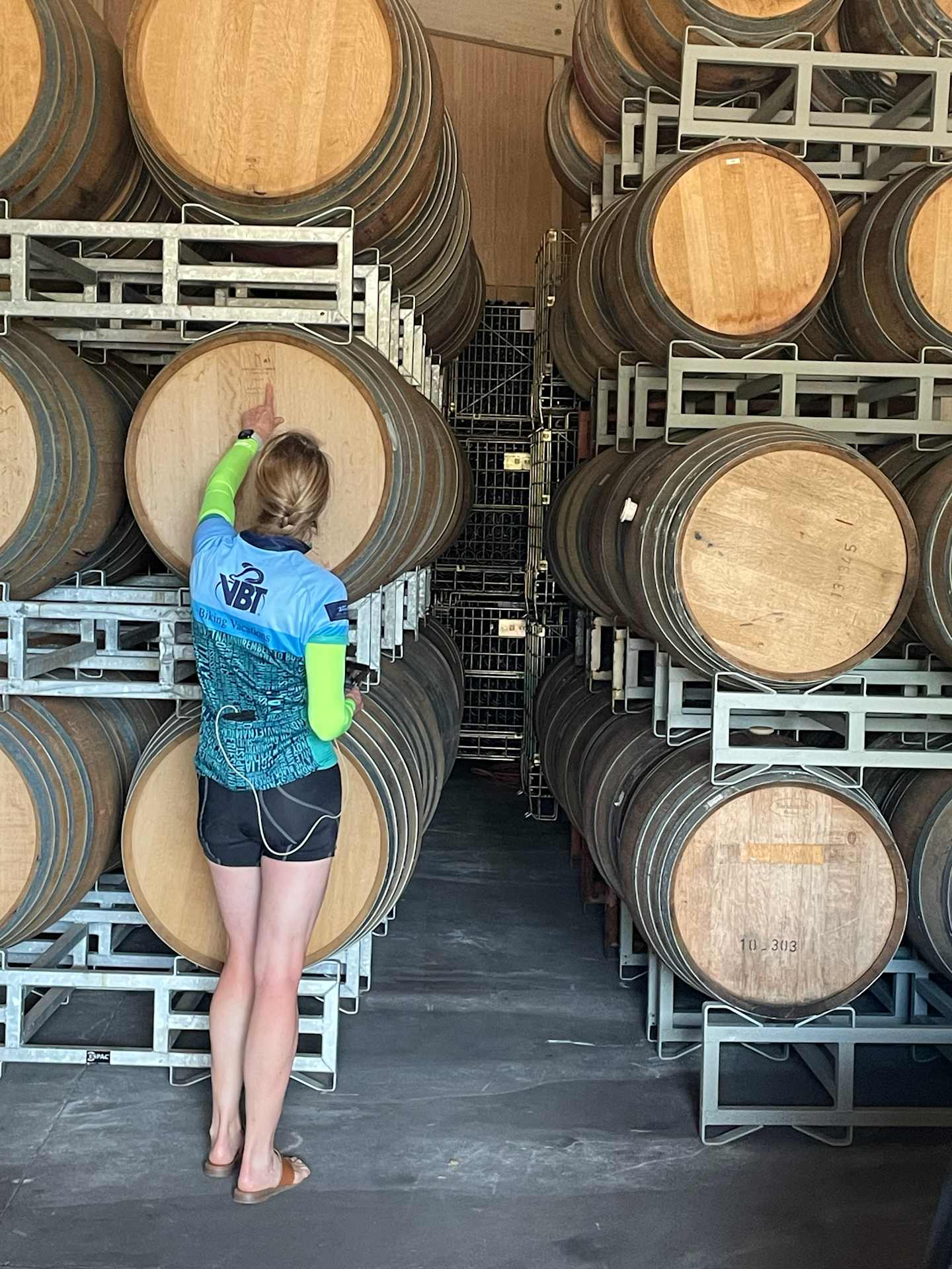 A person in a blue shirt and shorts is sitting among stacks of wooden barrels in a storage facility.