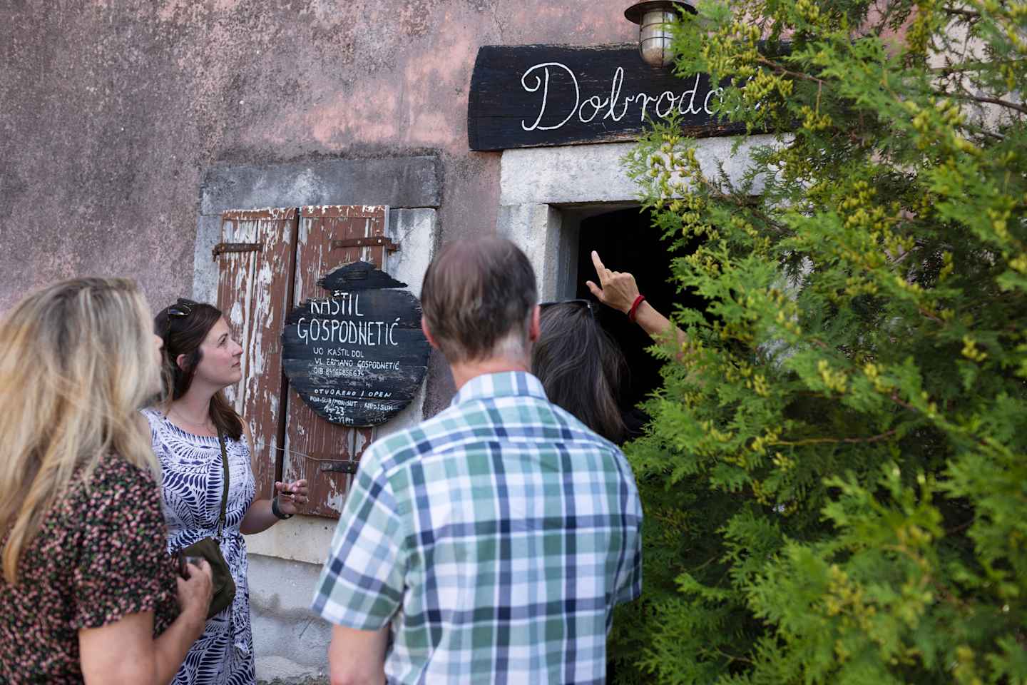 The image shows a group of people standing outside a building with a sign that reads "Delrado", with a lush green plant in the foreground.