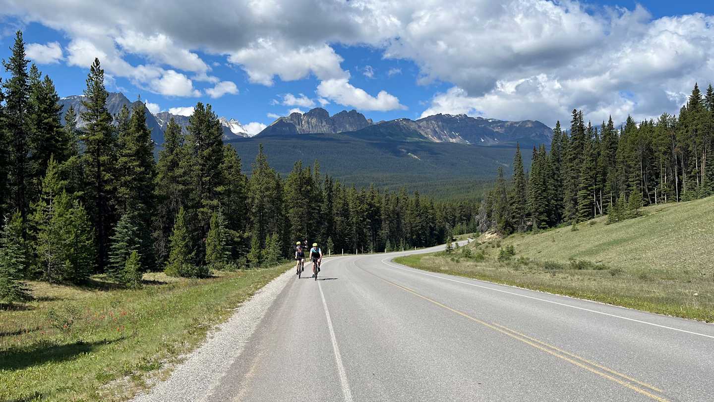 A winding road through a lush, forested landscape, with towering mountains visible in the distance against a cloudy blue sky.