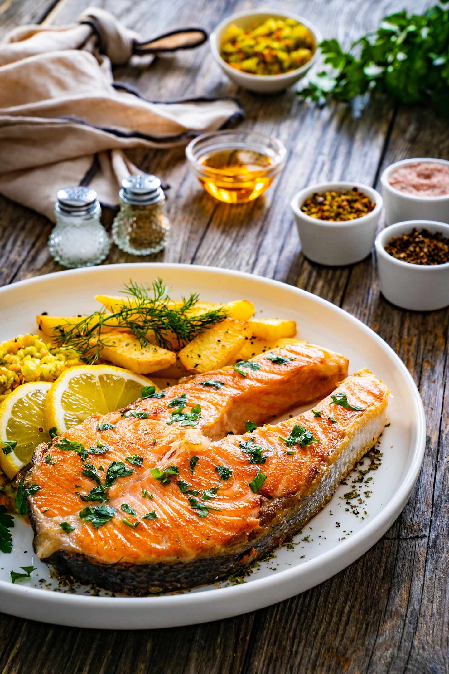 A plate of grilled salmon fillet served with lemon slices, herbs, and roasted potatoes on a wooden table, surrounded by various spices and condiments.