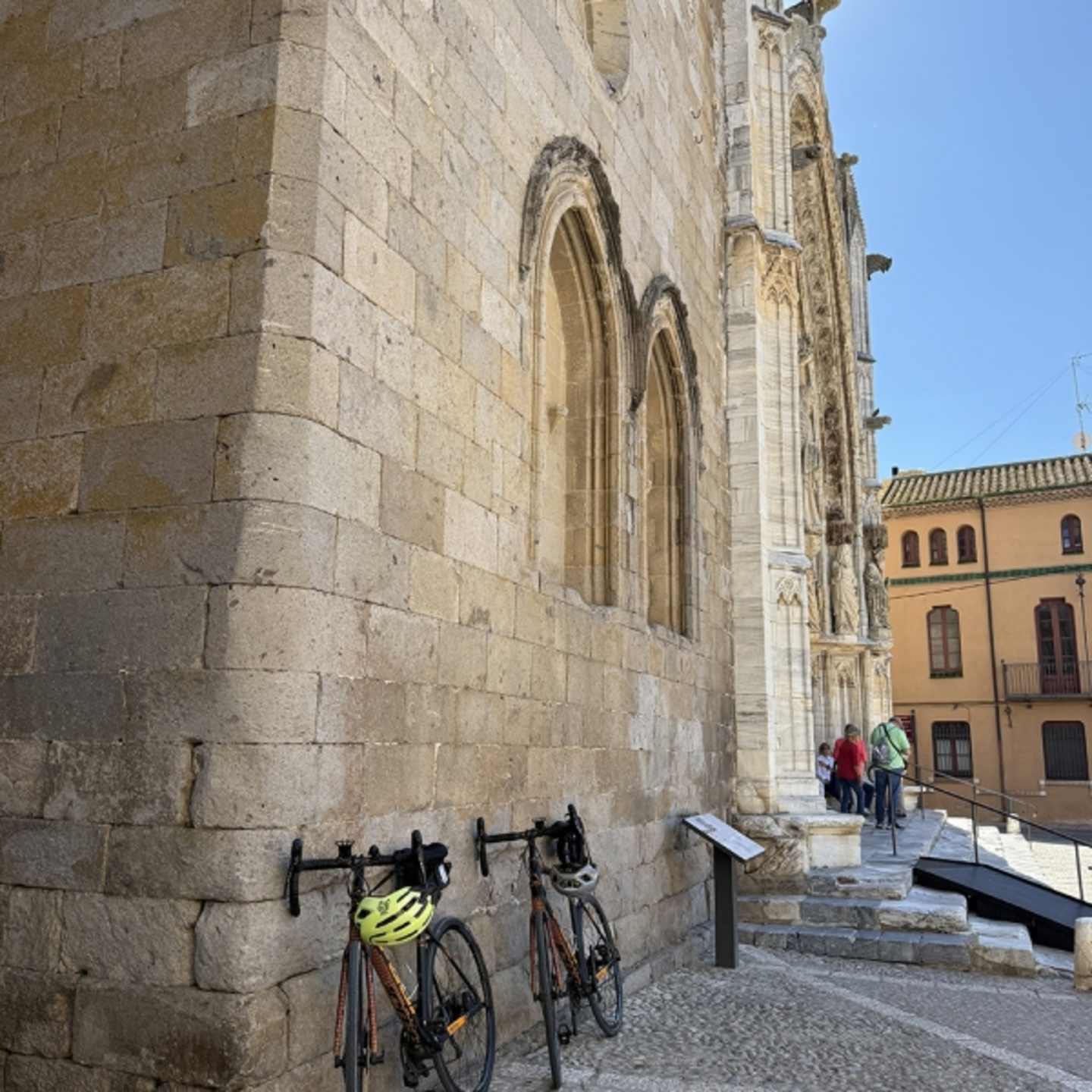 A historic stone building with ornate arched windows and doorways, with bicycles parked in the foreground and people walking on the steps leading up to the entrance.