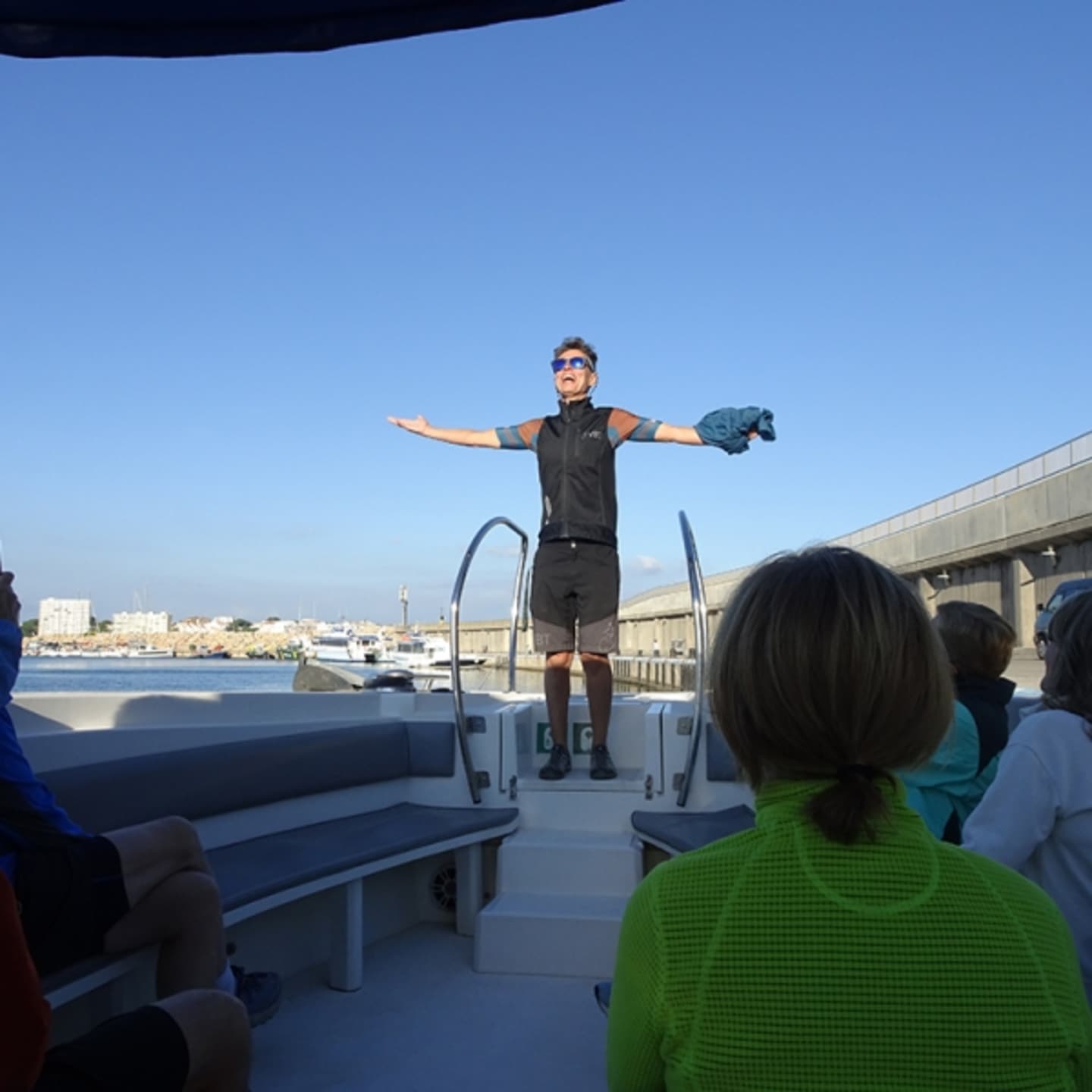 A person in a black outfit is standing on a boat deck, with a body of water and buildings in the background.