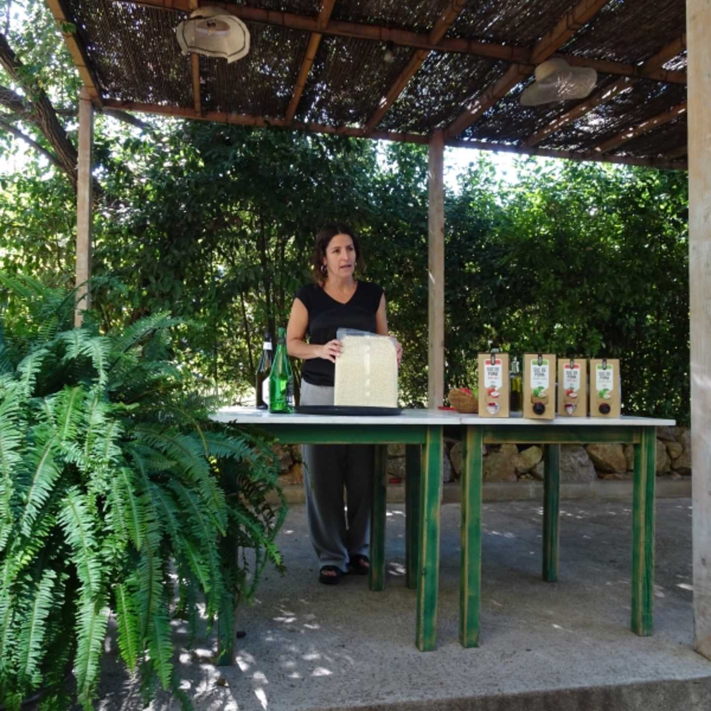A woman stands behind a table displaying various products in an outdoor setting surrounded by lush greenery and a thatched roof structure.