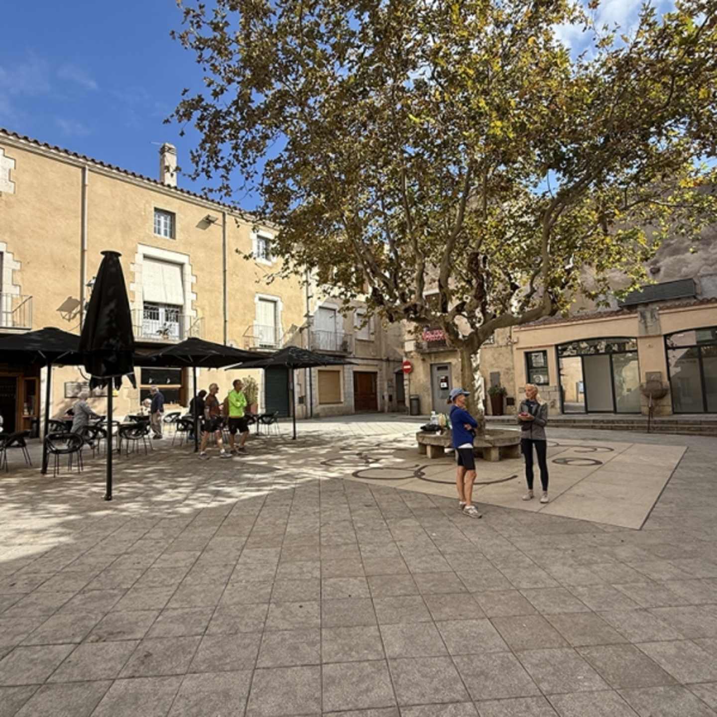 A bustling town square with a paved plaza, surrounded by buildings and trees, with people walking and enjoying the outdoor space.