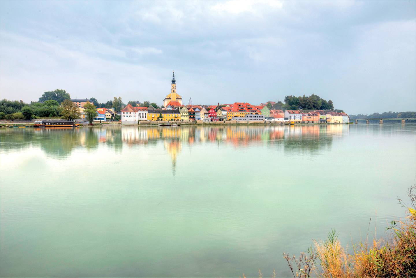 A picturesque town with colorful buildings and a church steeple reflected in the calm, turquoise waters of a lake or river in the foreground.