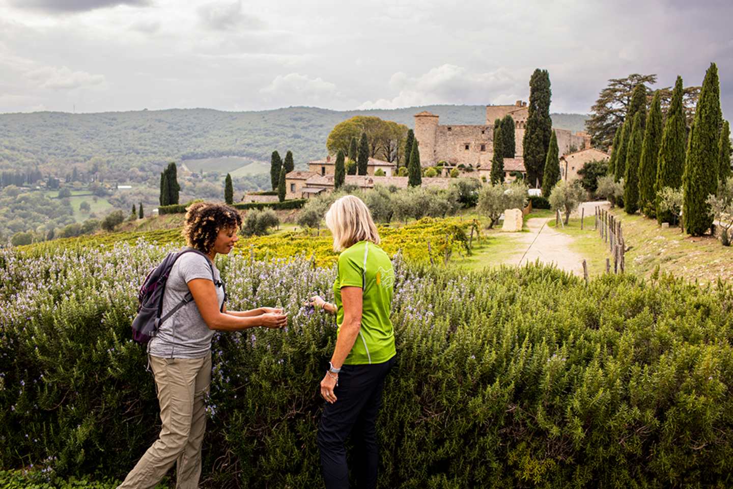 Two people, a man and a woman, are walking on a path through a lush, green landscape with a medieval-style castle or villa in the background, surrounded by cypress trees and rolling hills.