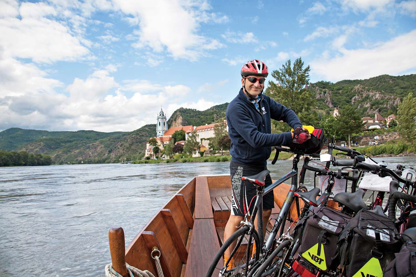 A cyclist stands on a wooden dock overlooking a scenic lake surrounded by mountains and a historic town in the background.