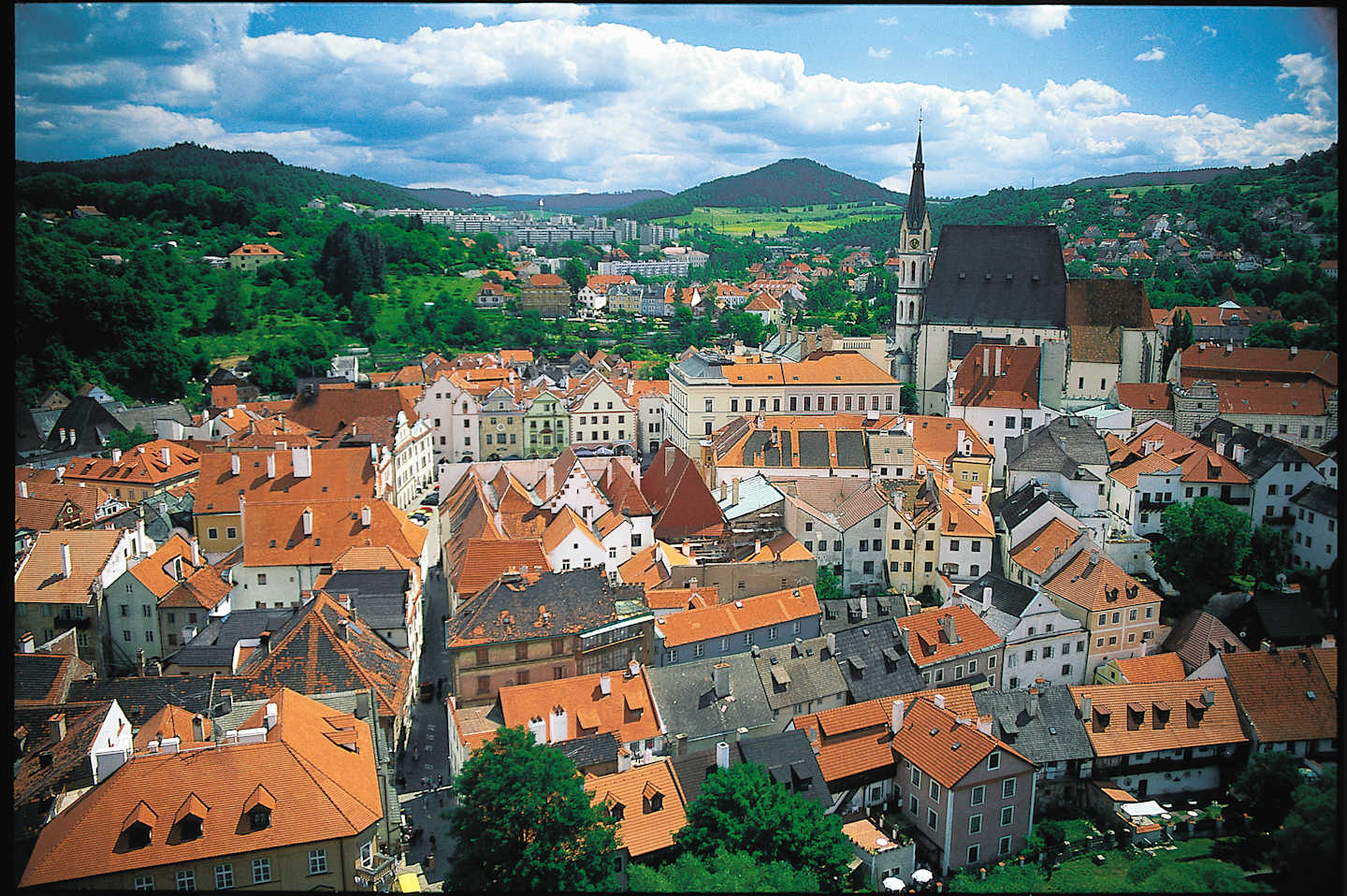 A picturesque European city nestled in a valley, with colorful rooftops, church spires, and lush green hills in the background under a cloudy blue sky.