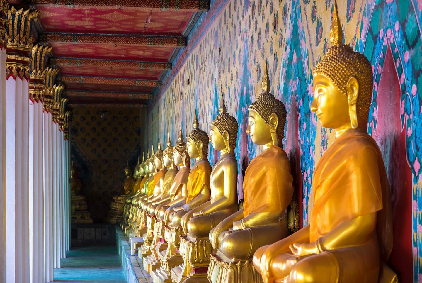 A long row of golden Buddha statues seated in a colorful, ornate temple hallway with intricate architectural details and vibrant wall paintings.