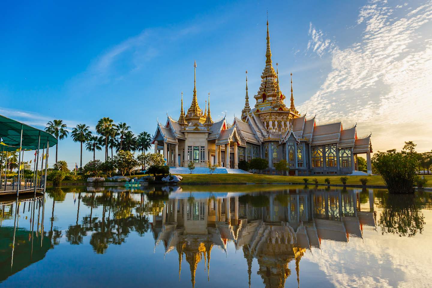 A magnificent Buddhist temple with ornate golden spires and intricate architectural details is reflected in a serene pond, surrounded by lush palm trees and a clear blue sky.