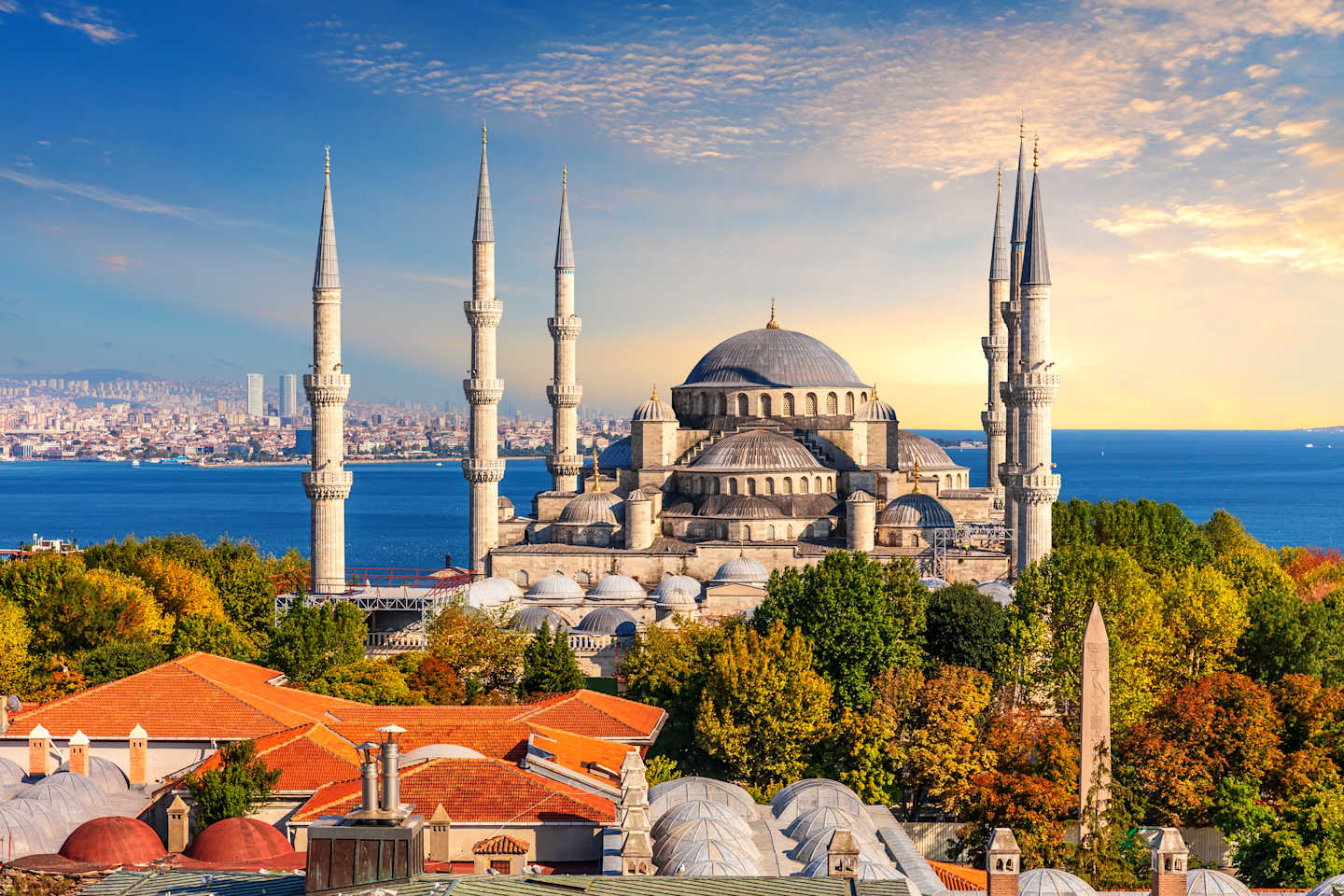 A magnificent mosque with multiple minarets stands prominently against a backdrop of a vibrant cityscape, surrounded by lush autumn foliage and a serene body of water.