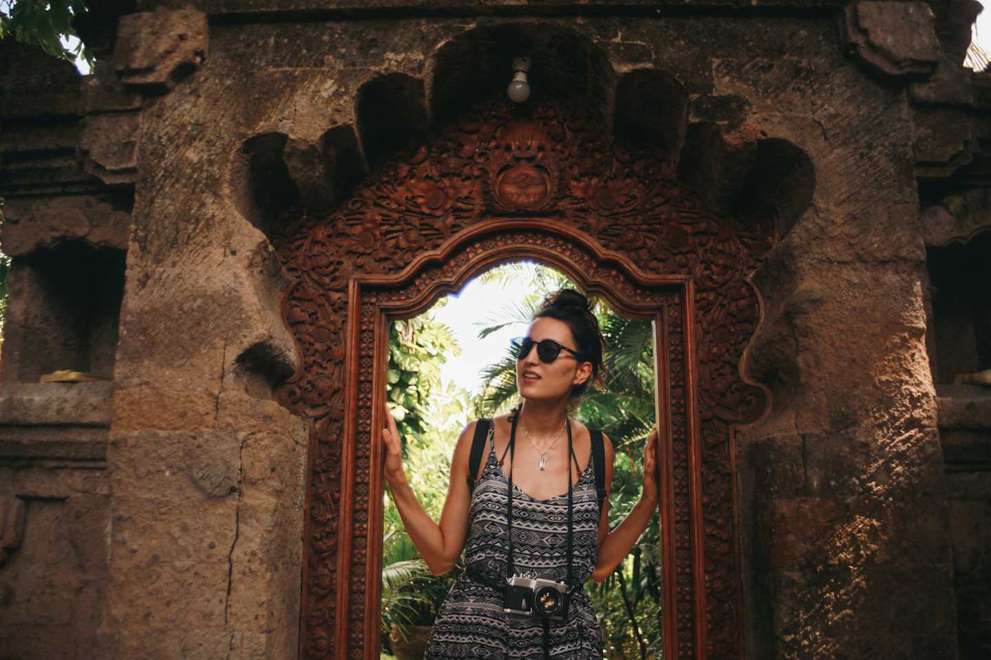 A woman in a patterned dress stands framed by an ornate stone archway, with lush greenery visible in the background.