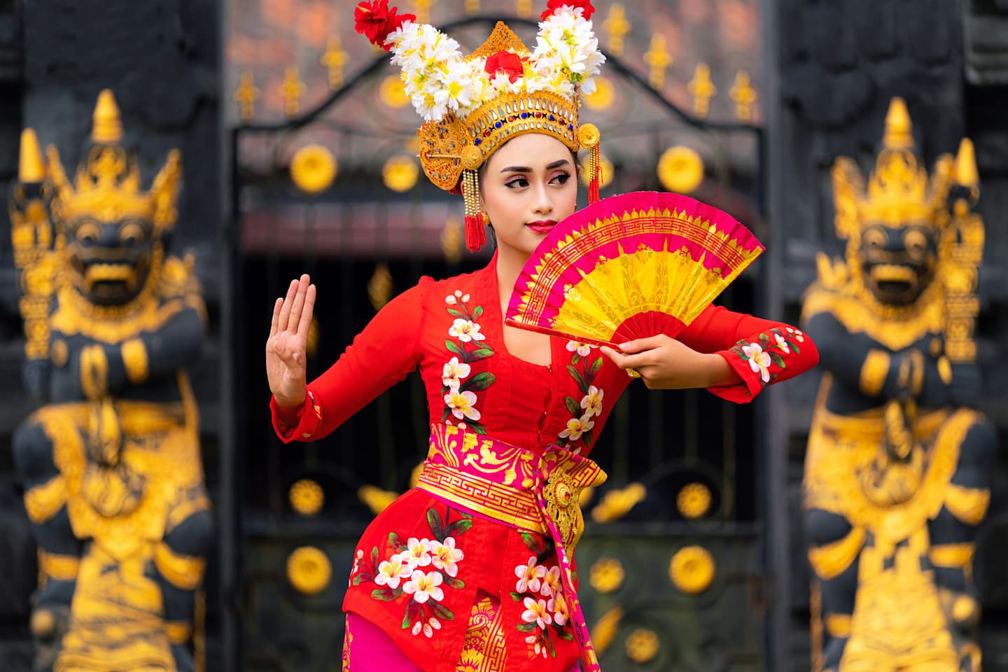 A woman in traditional Indonesian costume, wearing a vibrant red dress and headdress, holds a colorful fan and poses in front of ornate wooden carvings.