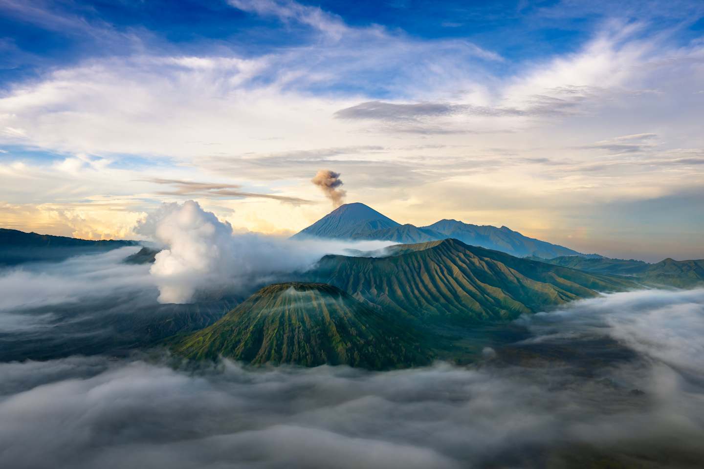 A majestic volcanic landscape emerges from the clouds, with a towering peak spewing smoke against a vibrant sky at sunset.