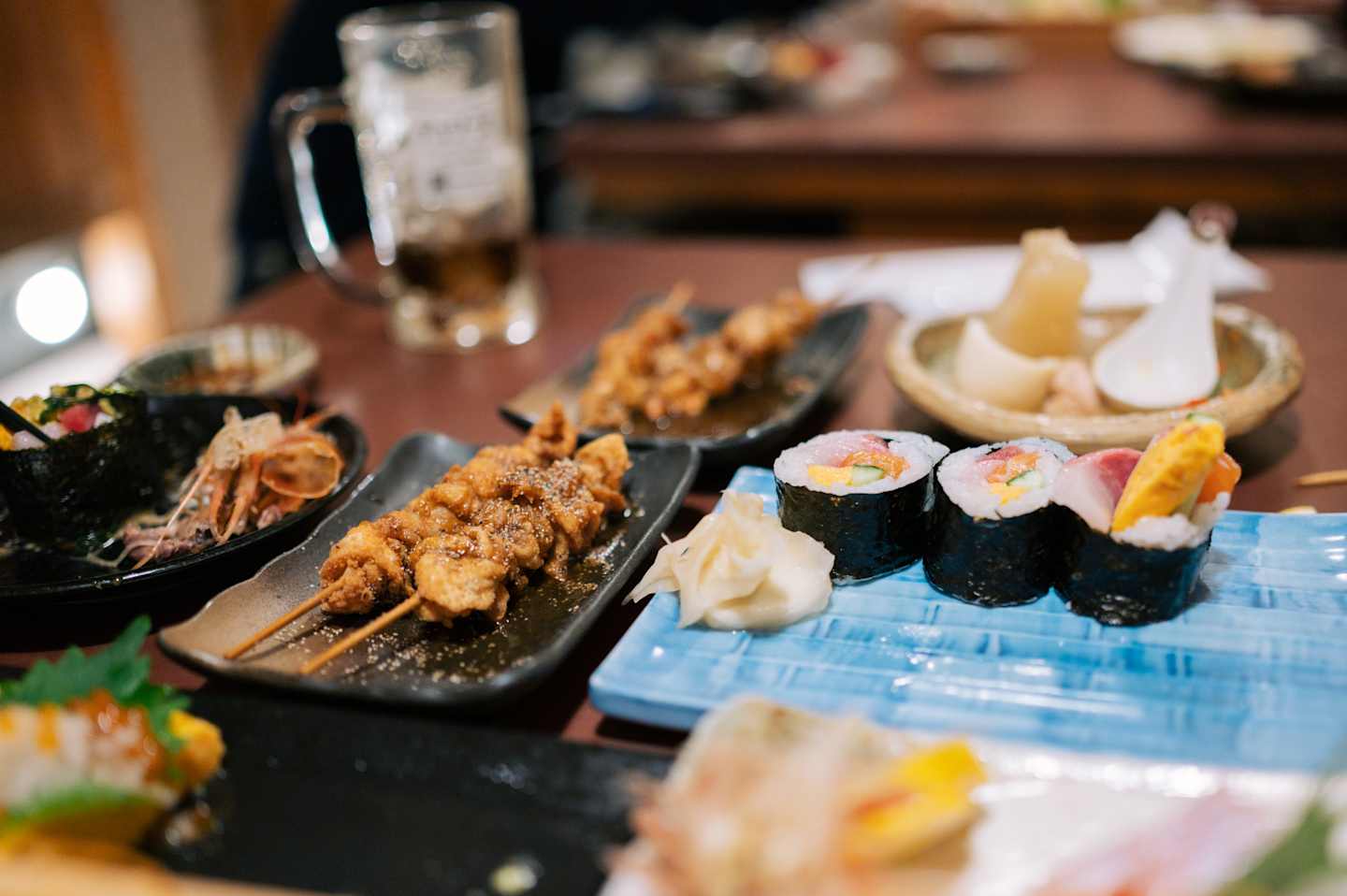 A variety of Japanese cuisine, including fried chicken, sushi rolls, and other dishes, is displayed on a wooden table with a blurred background.