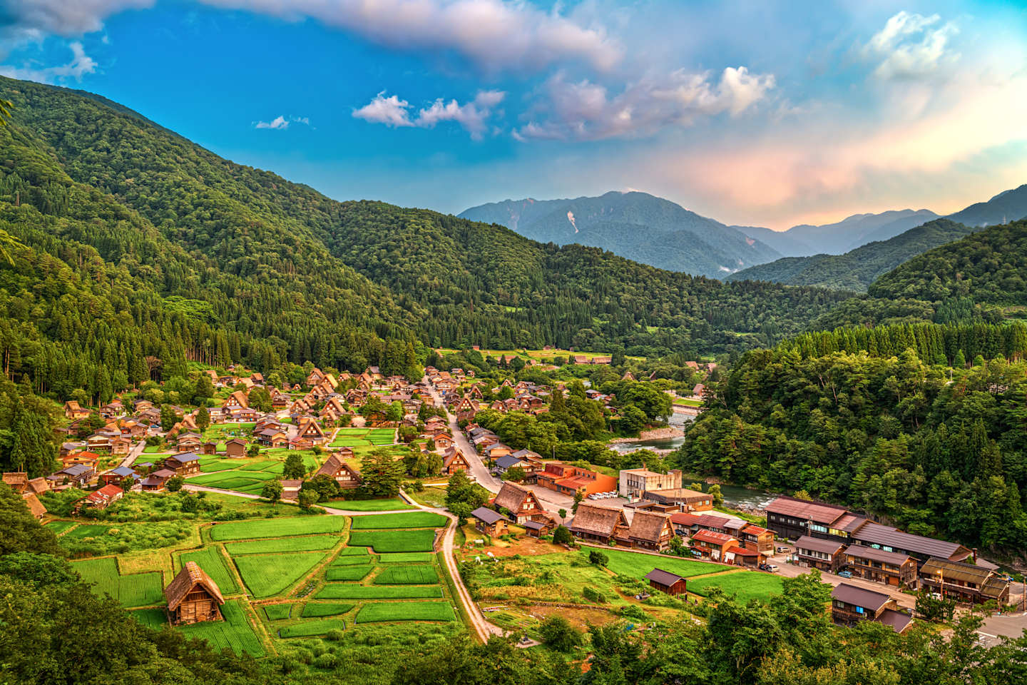 A picturesque village nestled amidst lush green hills and mountains, with colorful rooftops and fields stretching out in the foreground under a dramatic, cloudy sky.