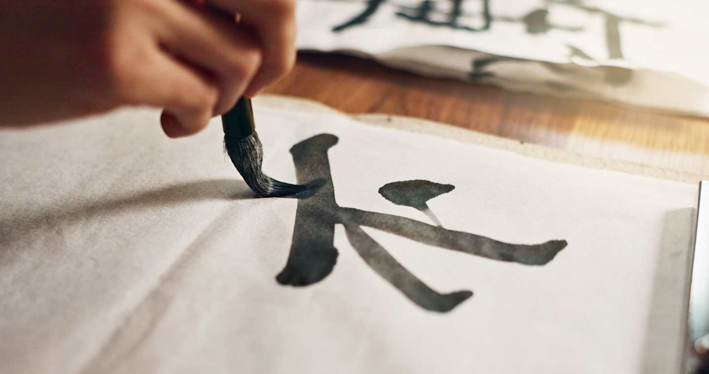 A hand is carefully brushing a calligraphic character onto a white surface, with blurred background elements suggesting a workspace or studio setting.