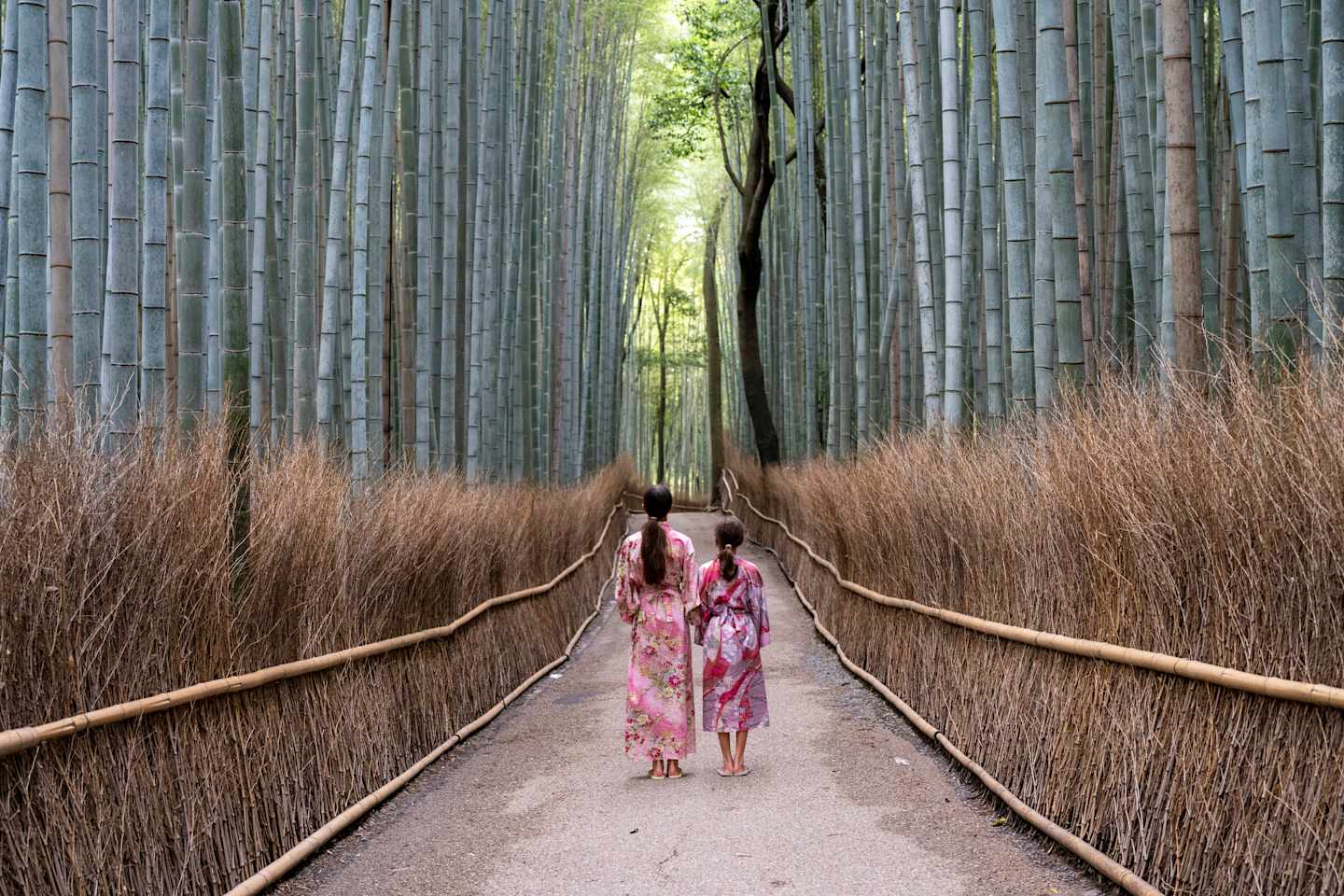 A winding wooden walkway leads through a serene bamboo forest, with a person in a colorful kimono walking along the path.