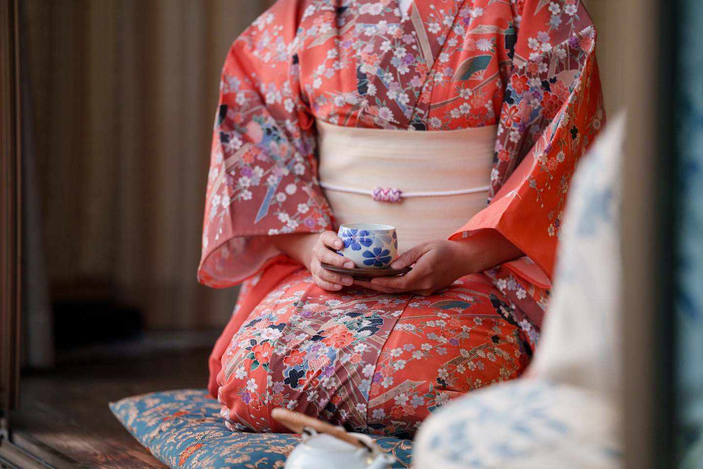 A person wearing a vibrant, floral-patterned kimono is holding a traditional Japanese teacup, seated against a backdrop of a dimly lit, textured wall.