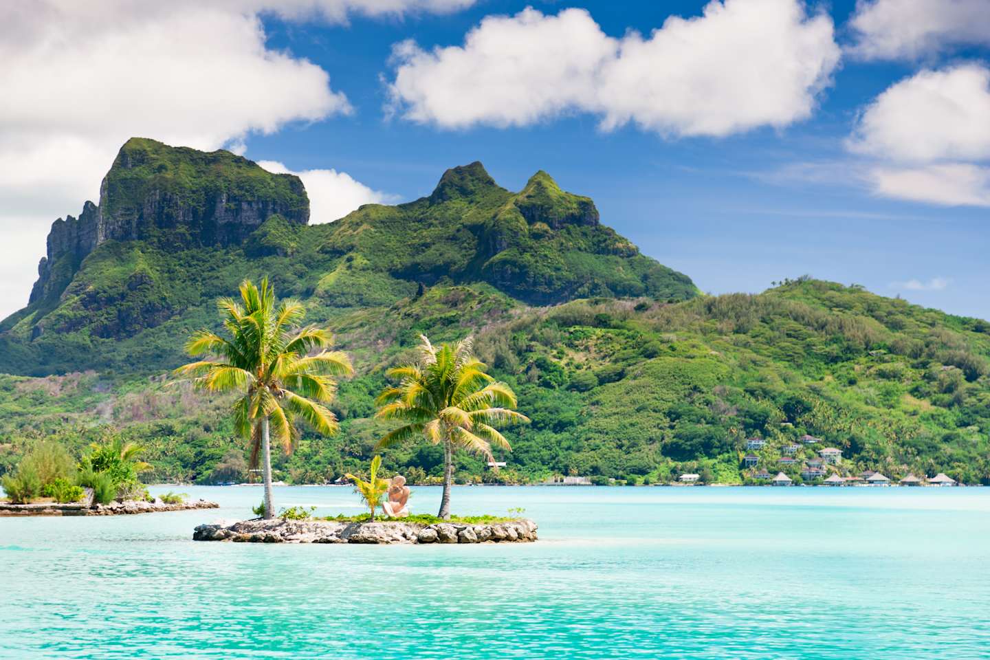 Lush, verdant mountains rise majestically from a tranquil, turquoise lagoon, with swaying palm trees adorning the foreground and a vibrant blue sky dotted with fluffy white clouds overhead.