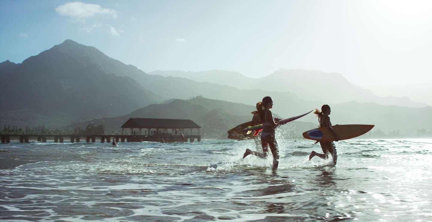 Two people with surfboards wading in the shallow waters of a tropical lagoon, with lush, mountainous terrain visible in the background.