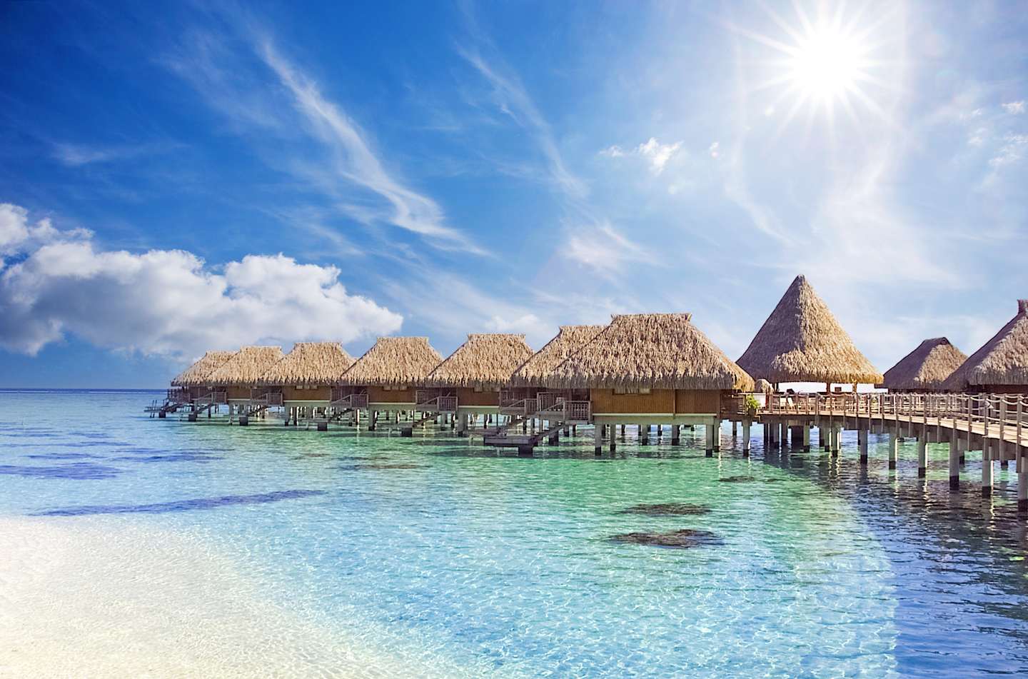 Overwater bungalows with thatched roofs sit atop a crystal-clear turquoise lagoon, surrounded by a vibrant blue sky dotted with wispy clouds and a shining sun.