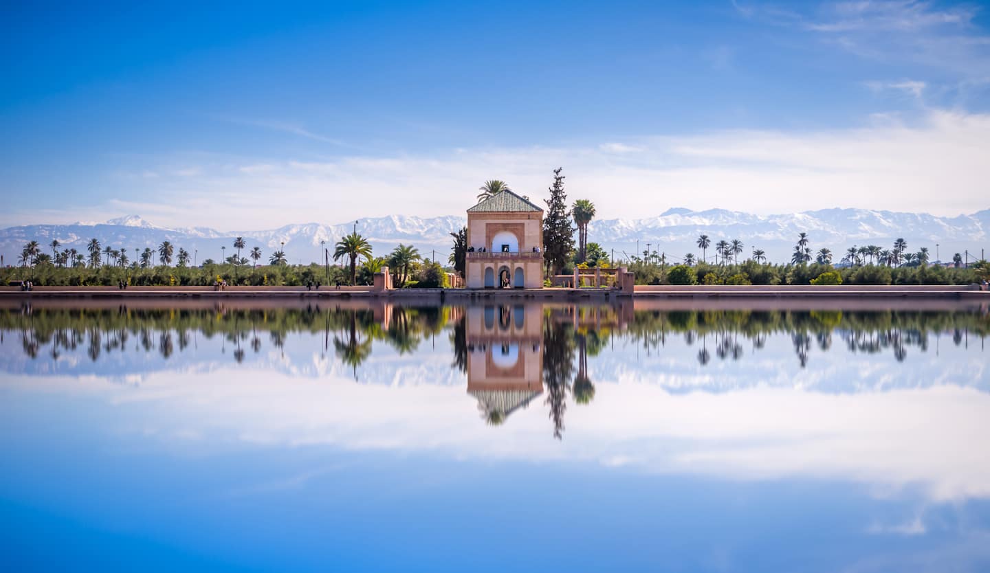 A serene and picturesque landscape with a pink-hued pavilion reflected in a tranquil body of water, surrounded by lush greenery and a clear blue sky.