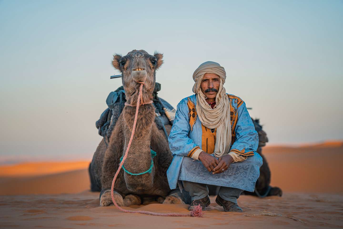 Two men, one riding a camel, stand in a desert landscape with a vibrant orange sky in the background.