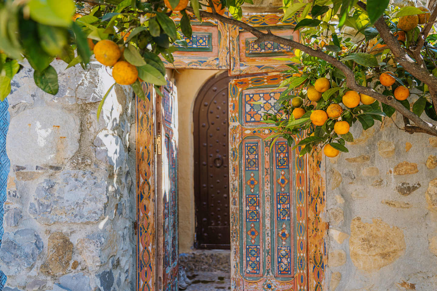 A rustic wooden door with intricate, colorful patterns is framed by a stone wall, and lush orange trees with ripe fruit hang overhead, creating a vibrant and inviting scene.