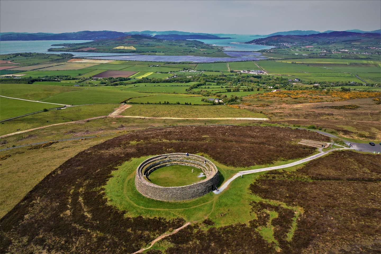 A circular stone fort sits on a grassy hill in the foreground, with a body of water and green fields stretching into the hazy background.