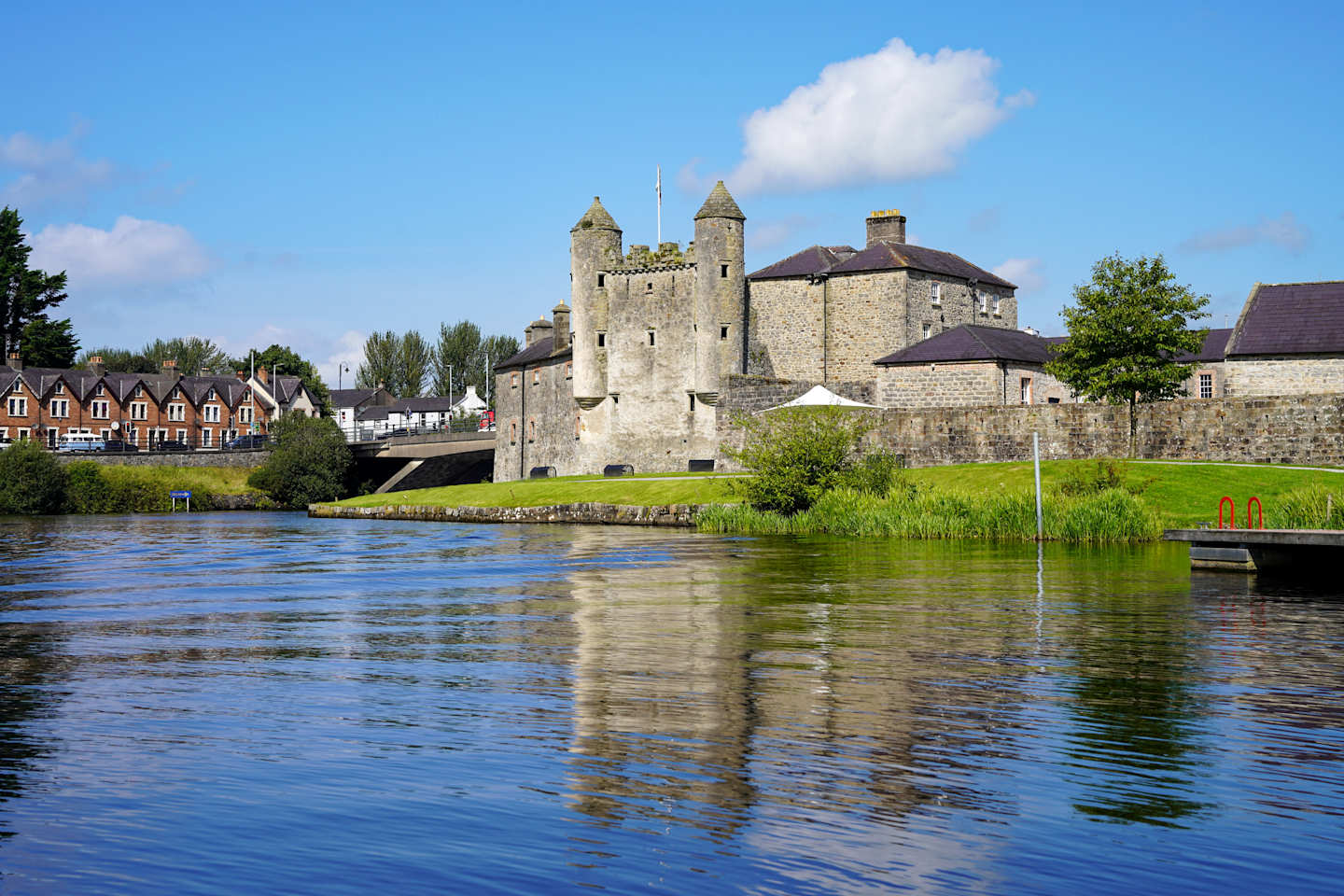 A wide, blue river flows in the foreground, reflecting the bright sky, while a stone castle with turrets stands prominently in the background, bordered by green grass and a few trees.