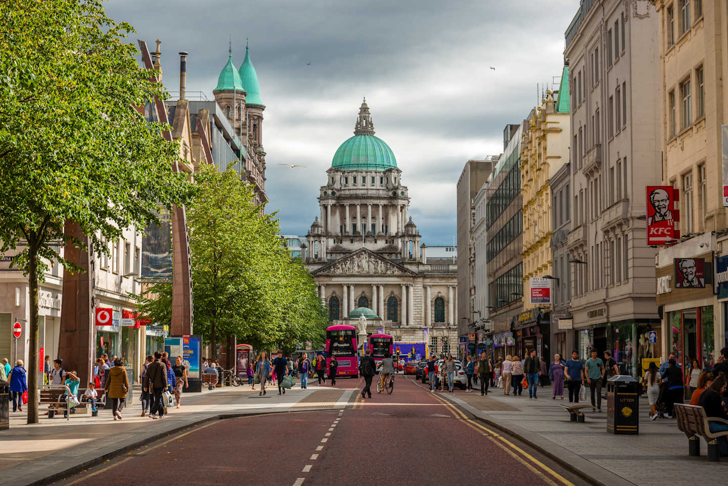 A bustling street with people and shops in the foreground leads to a grand domed building in the background.