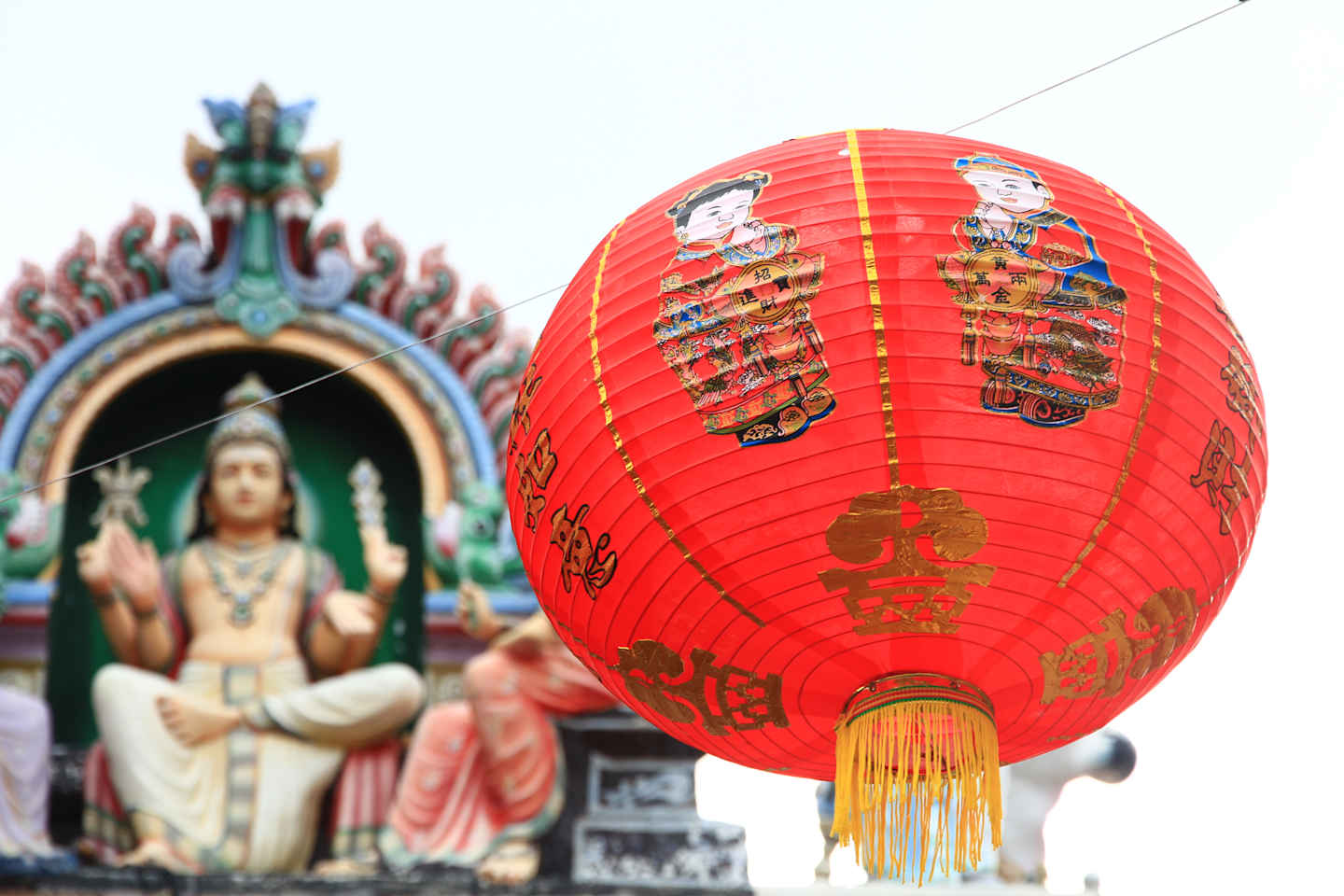 A bright red lantern with golden Chinese characters hangs in the foreground, obscuring a blurred background of a colorful Hindu temple with statues.