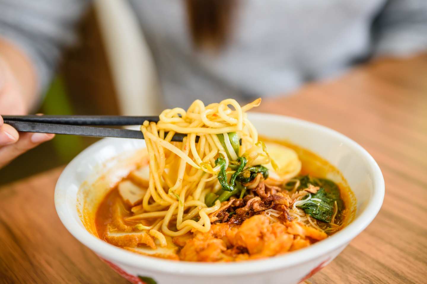 Chopsticks lift a portion of noodles from a steaming bowl of curry laksa, with a blurred person in the background.