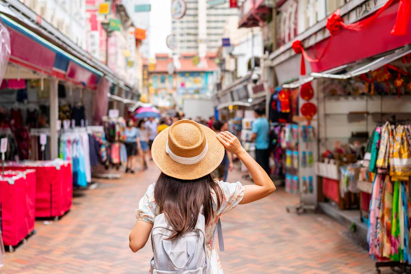 A woman in a straw hat and backpack walks down a bustling street market lined with shops and stalls, with buildings visible in the distance.