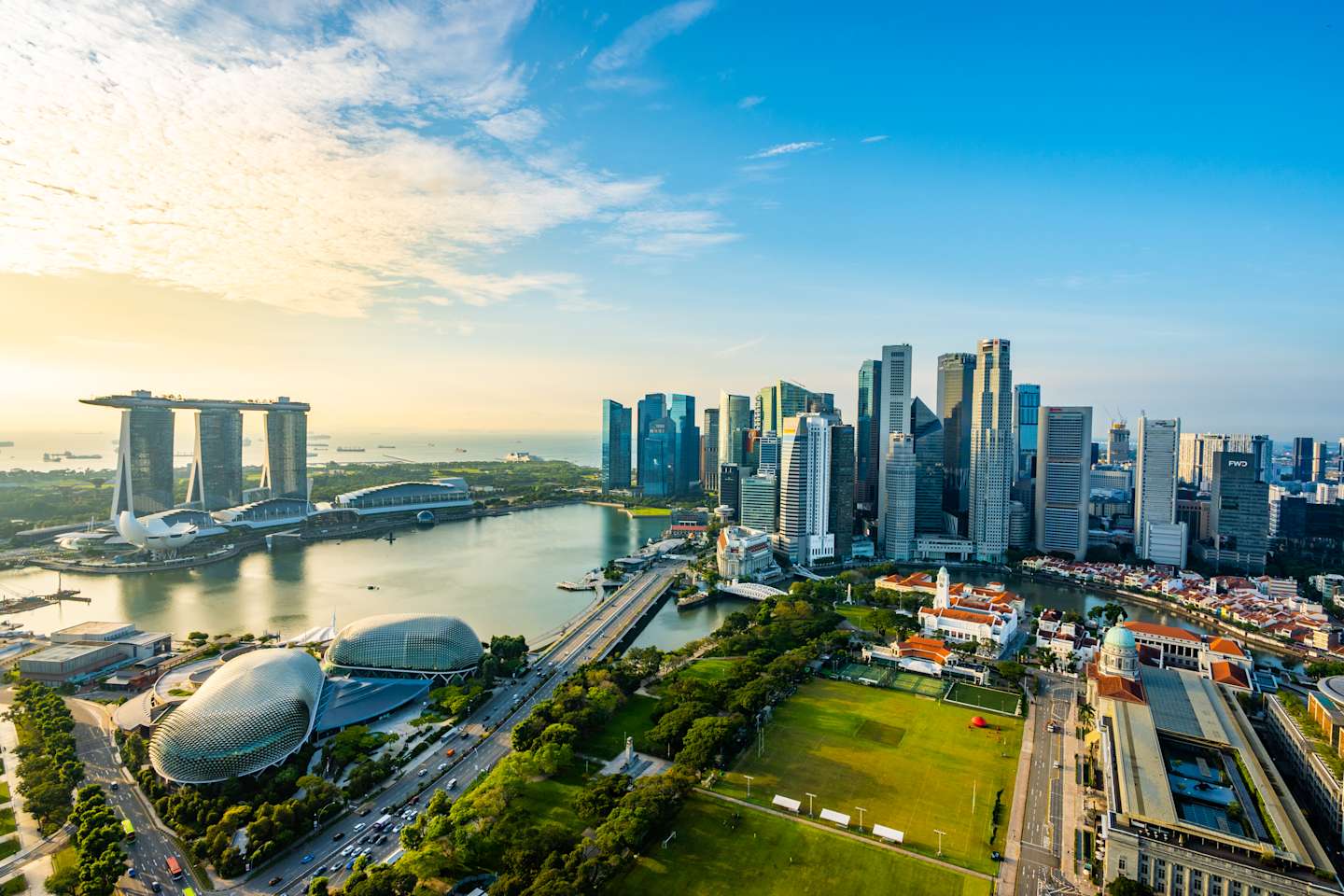 A vibrant cityscape with lush green parks and modern architecture in the foreground, and a bay dotted with iconic buildings and a sprawling skyline under a bright blue sky in the background.