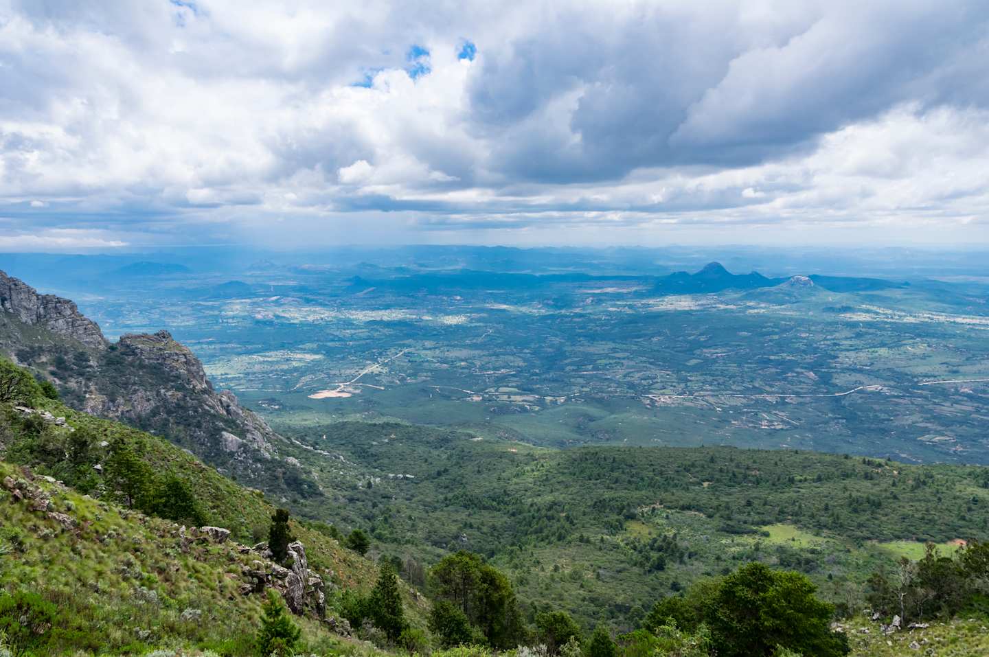 Green slopes and rocky outcrops in the foreground give way to a vast, hazy landscape dotted with distant mountains under a cloudy sky.