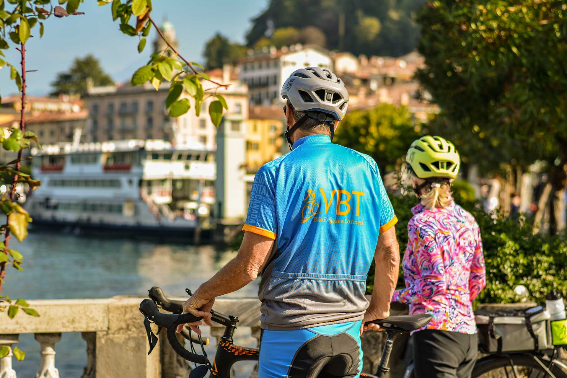 The image shows a person wearing a blue cycling jersey and helmet, standing in front of a scenic waterfront town with colorful buildings and trees.