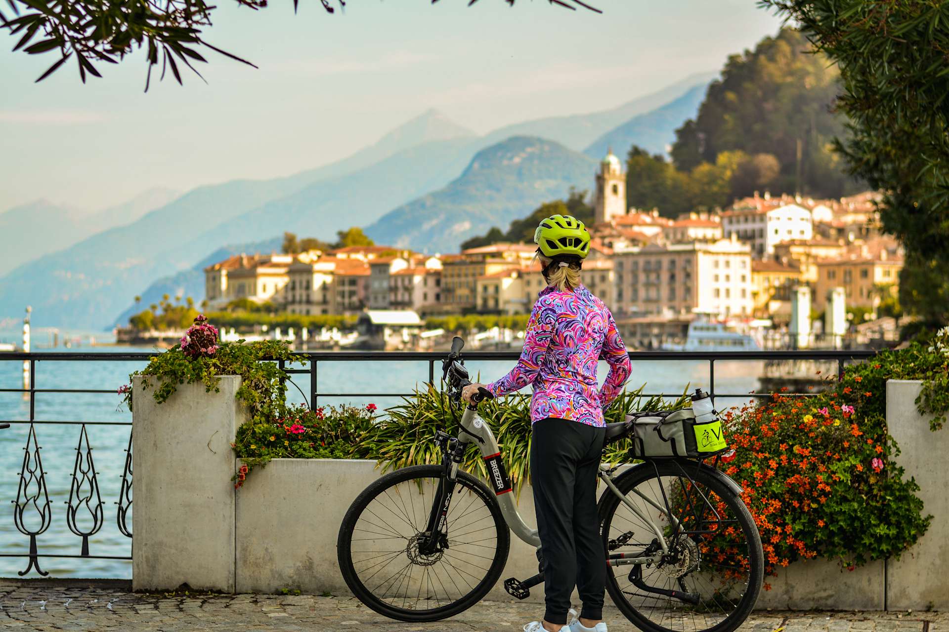 A person in a colorful floral jacket stands on a balcony overlooking a picturesque town nestled between mountains, with a bicycle in the foreground.